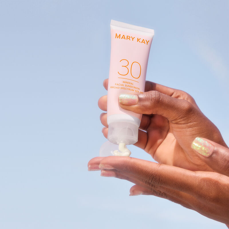 Pair of hands with deep skin tone and glitter nails squeezing product from a bottle of Mary Kay Mineral Facial Sunscreen Broad Spectrum SPF 30