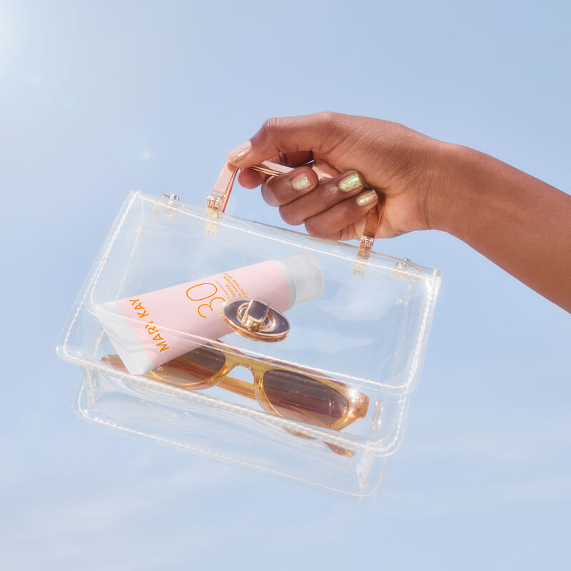 Hand with deep skin tone and glitter nails holding a small clear purse containing bottle of Mary Kay Mineral Facial Sunscreen Broad Spectrum SPF 30 an