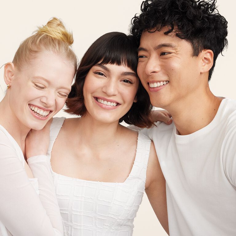 Dos mujeres y un hombre de pie, muy juntos y con los brazos alrededor del otro, vistiendo camisas blancas. Mary Kay.