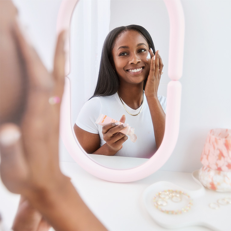 Mujer mir&aacute;ndose en un espejo de maquillaje y aplicando Protector Solar Facial Mineral FPS 30 Mary Kay en su mejilla.