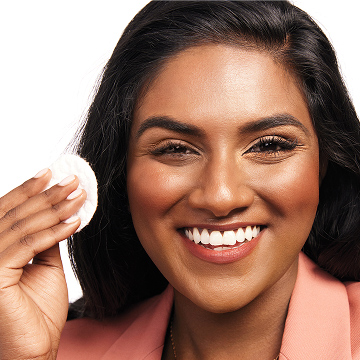 Modelo sonriendo a c&aacute;mara mientras se prepara para retirar el maquillaje de ojos con un disco desmaquillante.